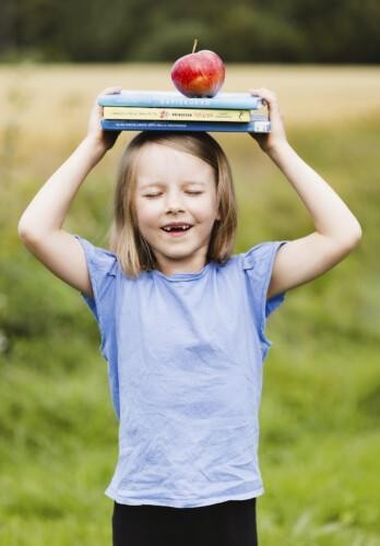 Tyttö pitää keskittyneesti kiinni päänsä päällä olevaa kolme kirjaa ja omenaa. Pellon laidalla kuvatun tytön hymy paljastaa edessä vajaan hammasrivistön. Maitohampaat ovat lähteneet.