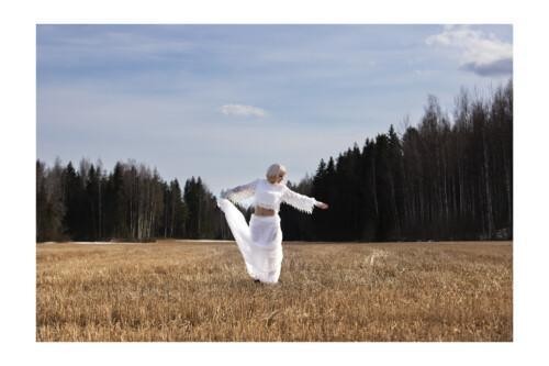Valkoisiin, läpikuultaviin pukeutunut nainen seisoo keskellä puitua peltoa, taustanaan metsikkö ja vaalea pilvinen taivas.