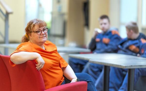 Rehtori Anne Laine istuu aulan sohvalla taustalla kone-ja tuotantotekniikan opsikelijoita.
